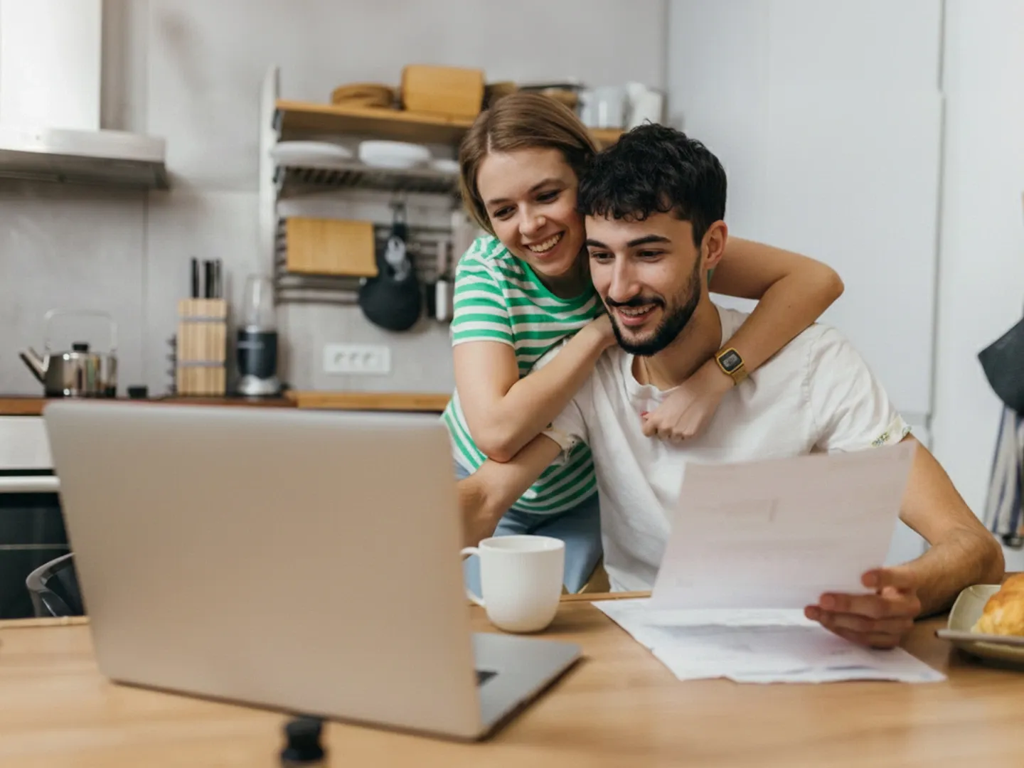 A smiling young couple looks at a laptop together while holding paperwork at a kitchen table.